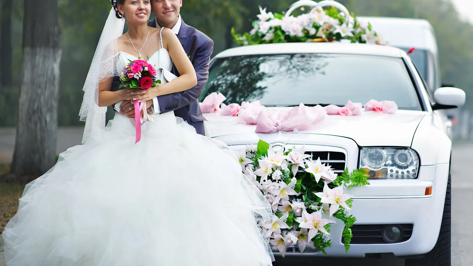 Bride and groom beside a white wedding car with flowers, chauffeur waiting