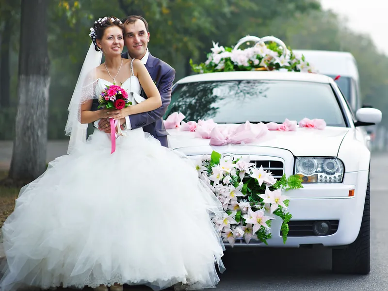 A bride and groom beside a white wedding car decorated with flowers as the chauffeur waits